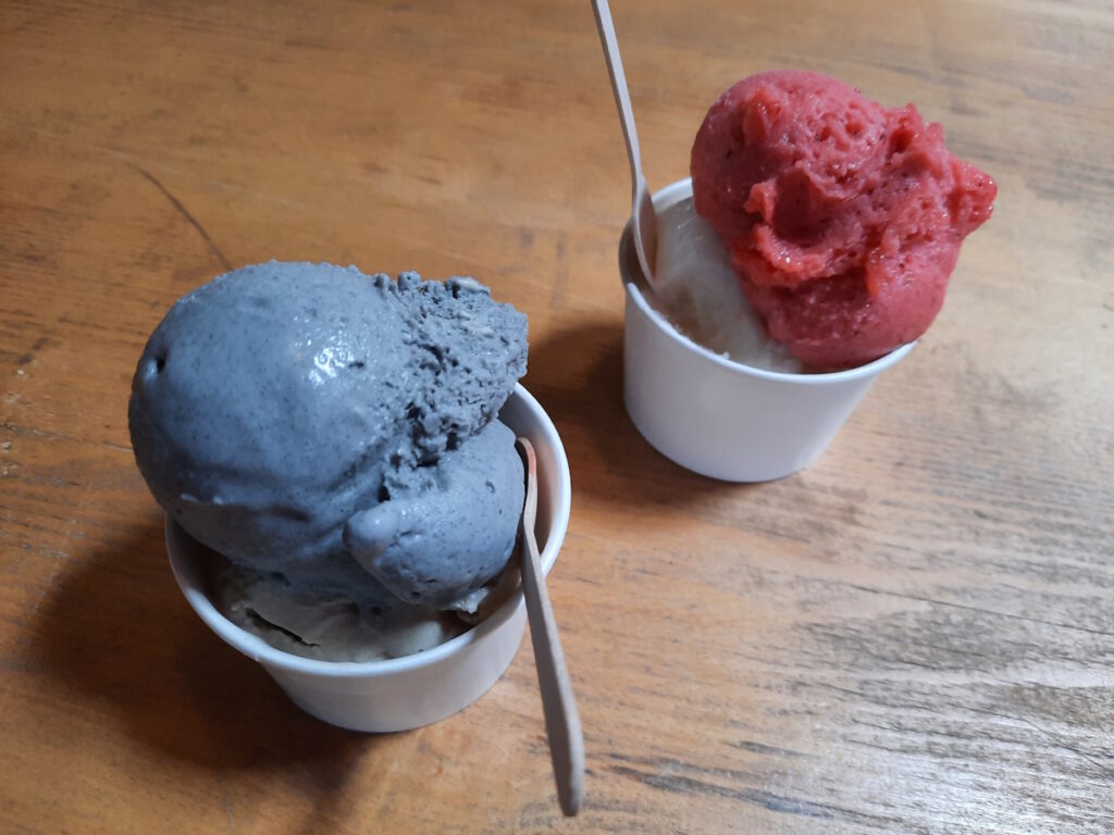 Zwei Pappbecher mit Eiskugeln. Vorn eine gräuliche Kugel Schwarzes-Sesam-Eis, hinten eine dunkelrote Kugel Erdbeereis.