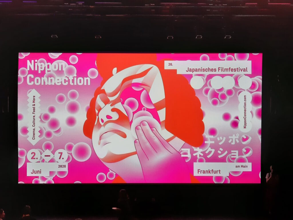 Das diesjährige Logo der Nippon Connection auf einer Filmleinwand. Es zeigt das Gesicht einer gezeichnete Kabuki-Figur und eine Hand mit einem Makeup-Pad. Ein Streifen des Gesichts wurde von dem MakeUp-Pad entfernt und zeigt den gemusterten Hintergrund des Logos.