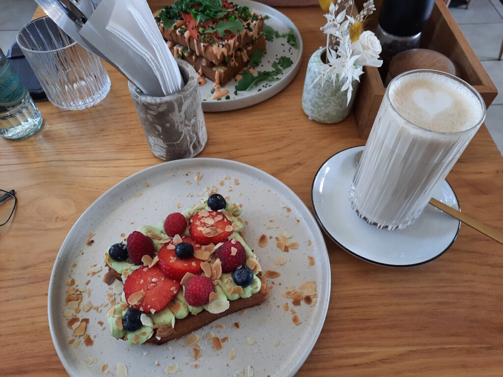 Zwei Teller mit unterschiedlichen Sandwiches. Auf meinem Teller liegt eine dicke Weißbrotscheibe mit Matcha-Creme, verschiedenen Beeren und Mandelscheibchen. Daneben ein Glas mit Chai Latte.