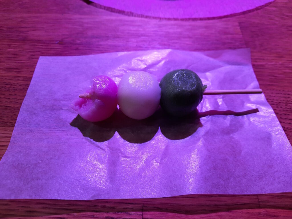 Klassisches Dango auf einem Papier liegend. Die rosa Mochi-Kugel ist schon angebissen.