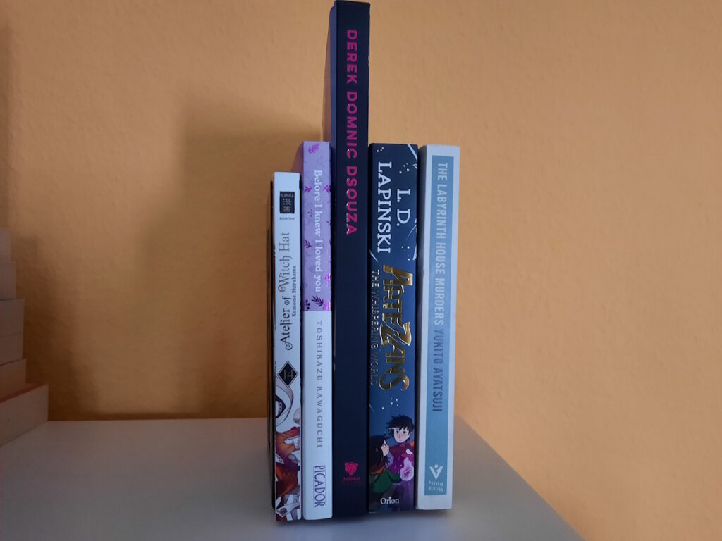 Ein paar Bücher und Manga zwischen zwei Buchstützen. Die Titel lauten von links nach rechts: Atelier of Witch Hat 14, Before I Knew I Loved You, The Art of Derek Domnik Dsouza, The Artezans - The Whispering World und The Labyrinth House Murders