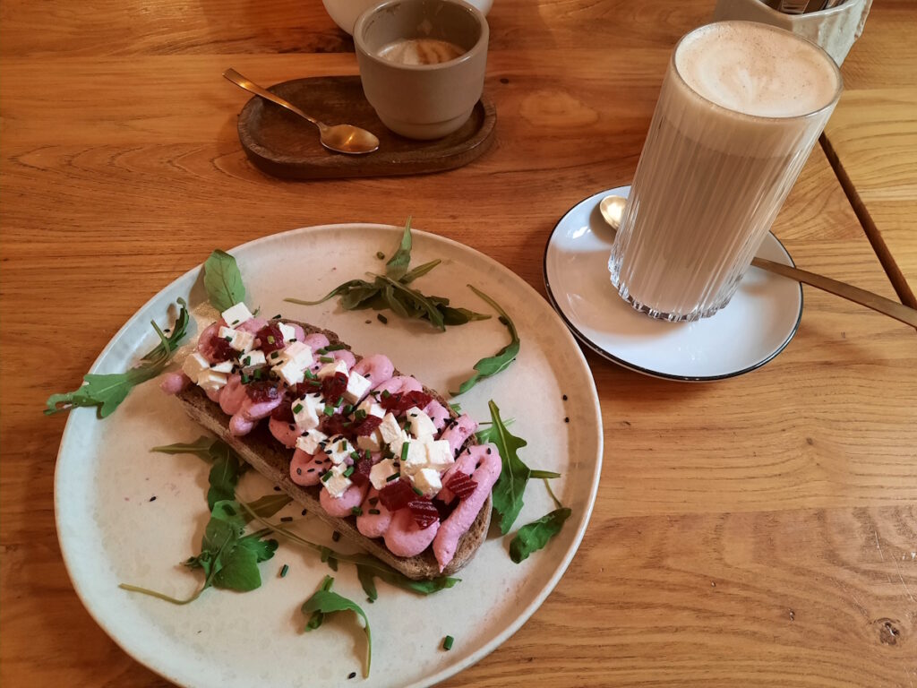 Ein Teller mit einem Brot mit Rote-Beete-Creme, Rote Beete Würfeln und weißem Käse. Daneben ein Glas mit Chai Latte.
