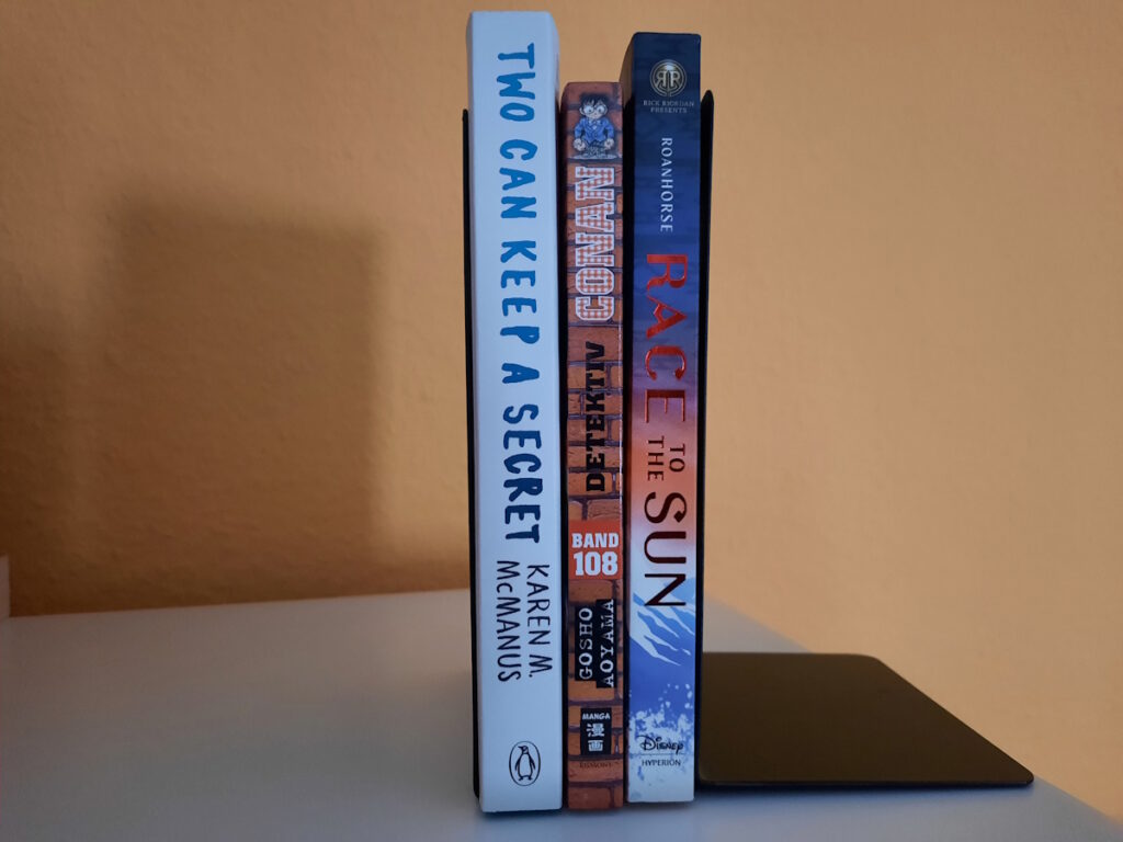 Drei Bücher zwischen zwei Buchstützen. Die Titel lauten von links nach rechts: Two Can Keep a Secret, Detektiv Conan 108 und Race to the Sun.