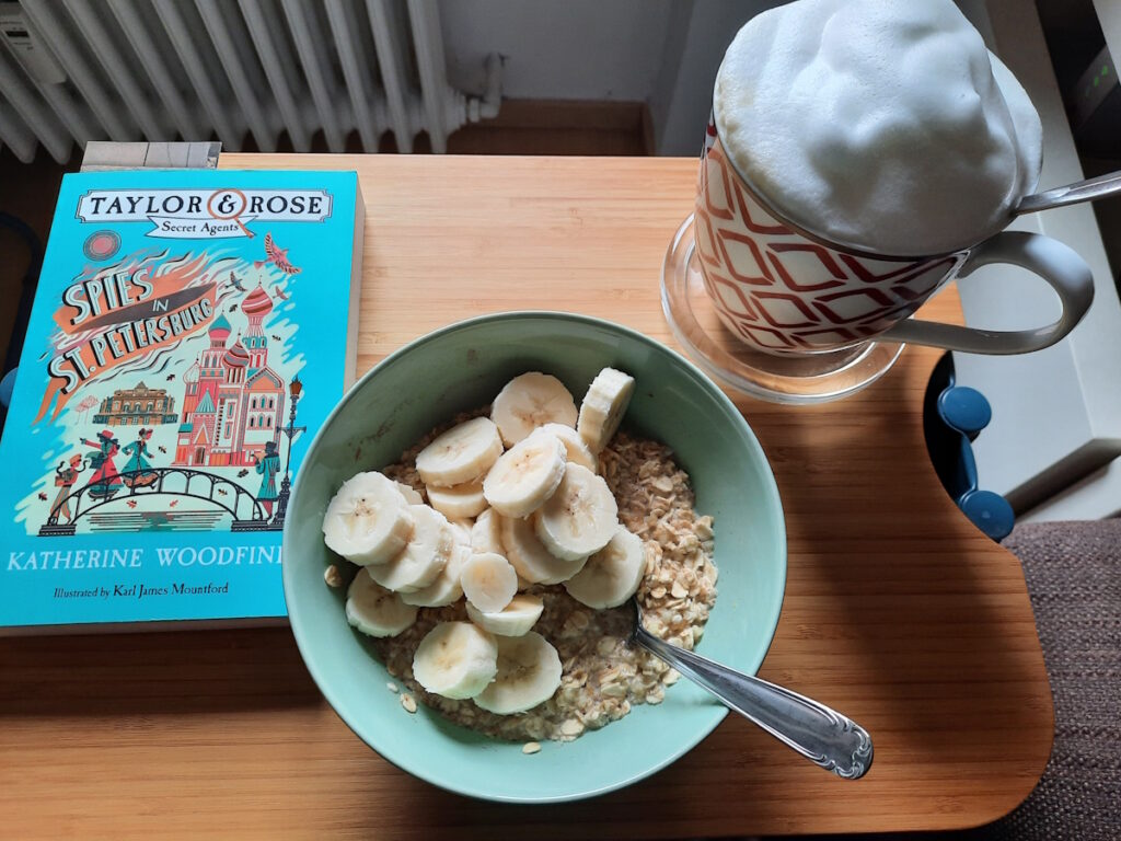 Eine hellgrüne Schale mit Porridge und Bananenscheiben. Daneben eine große Tasse mit Milchkaffee und einer üppigen Milchschaumhaube. Links von der Schale liegt noch die Taschenbuchausgabe von Spies in St. Petersburg, deren Cover einige Figuren auf einer stilisierten Brücke zeigt.
