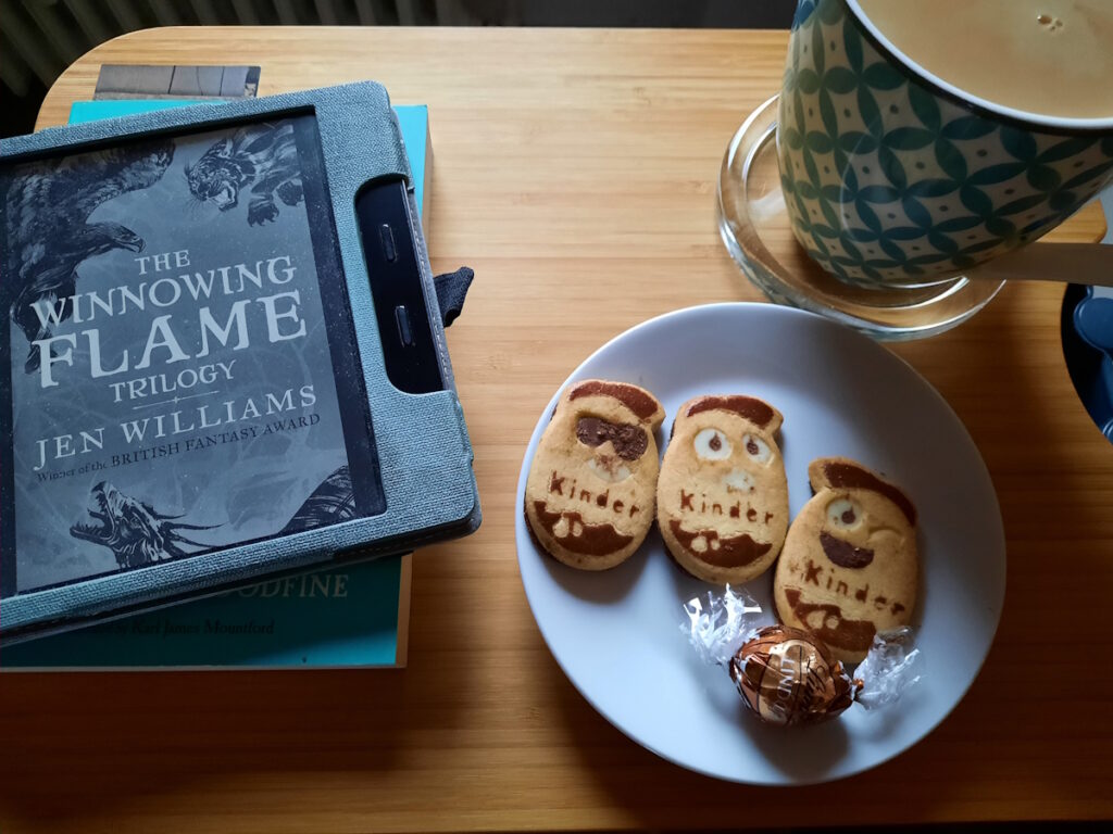 Ein kleiner Tisch mit einer Untertasse auf der drei Kekse und eine Schokokugel liegen. Im Hintergrund ist eine dampfende Tasse Tee zu erahnen und links vom Teller ein angeschalteter eReader mit dem Cover der "The Winnowing Flame"-Trilogie von Jen Williams.