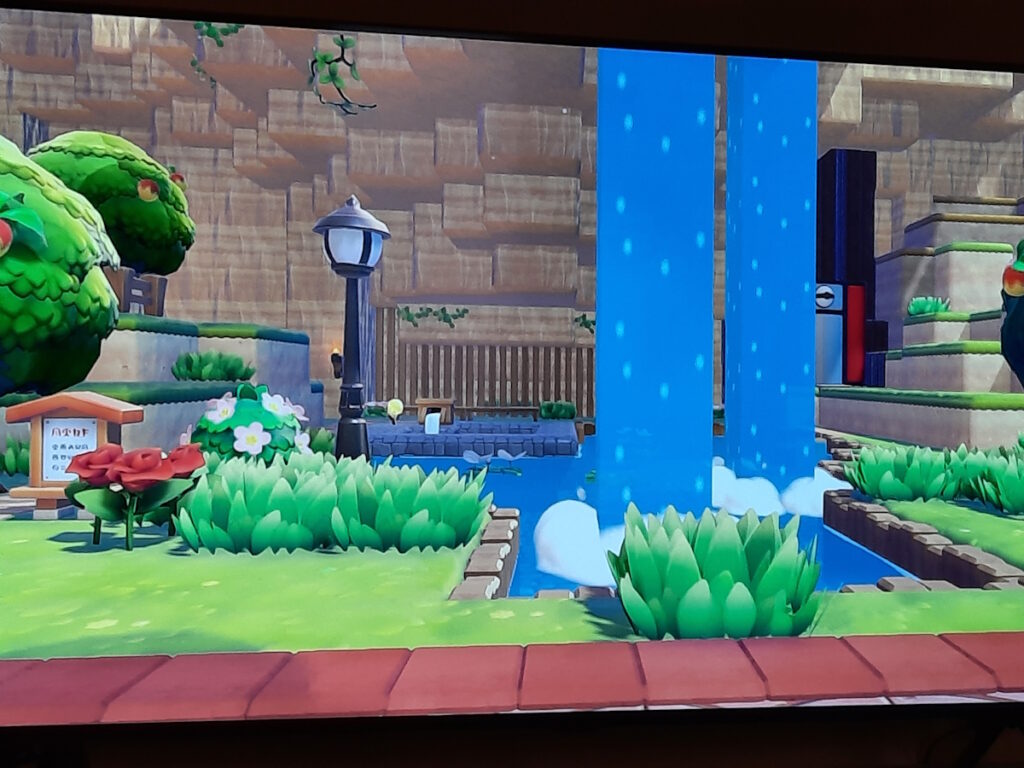 Ein doppelter Wasserfall in dem Spiel Pokopia, der in ein umrandetes Becken fällt. Im Hintergrund ist ein gemauerter Bereich mit dampfendem Wasser zu sehen, im Vordergrund eine altmodische Straßenlaterne.