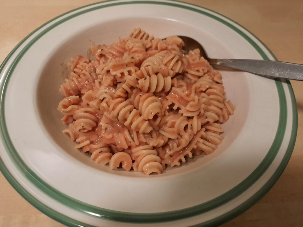 Ein cremefarbender, tiefer Tellermit zwei grünen Streifen am Rand. Im Teller befindet sich eine Portion Nudeln mit Basilikum-Tomaten-Sauce.