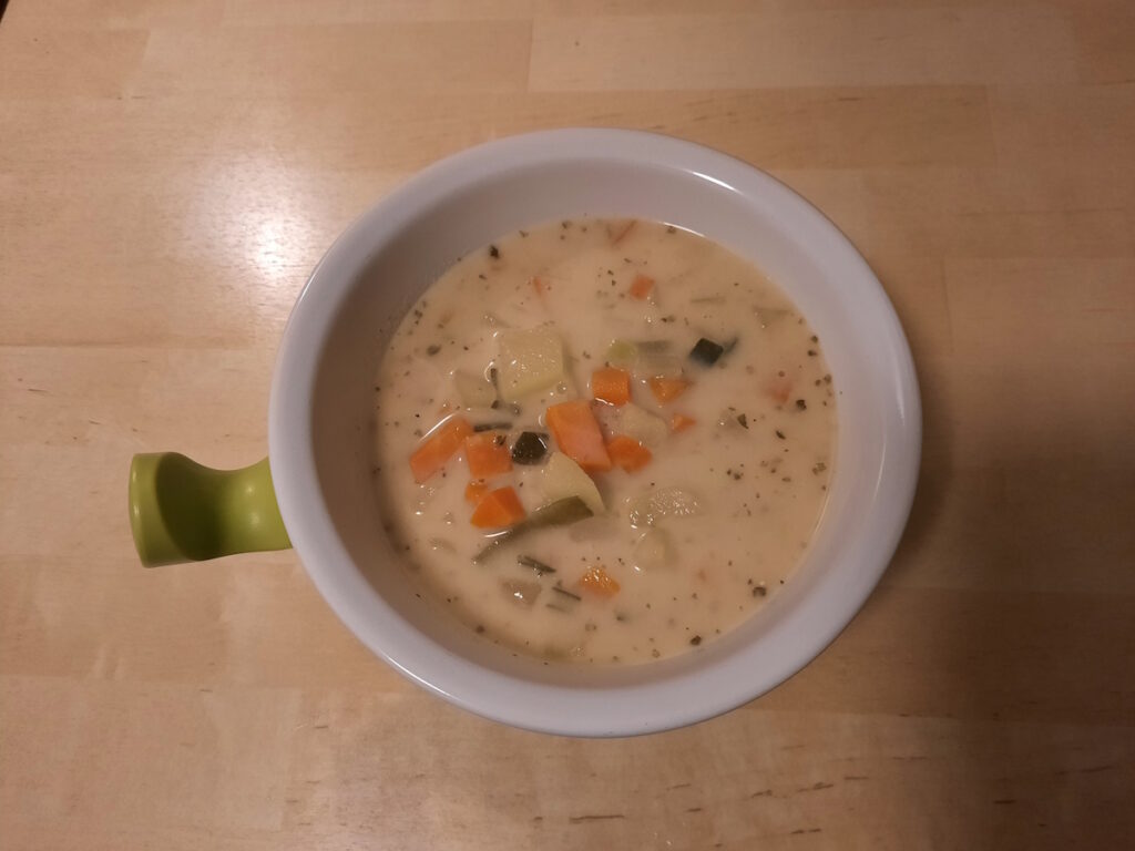 Eine Schale mit cremiger Kartoffelsuppe, in der Möhren- und Lauchstückchen, sowie einige Kräuter schwimmen.