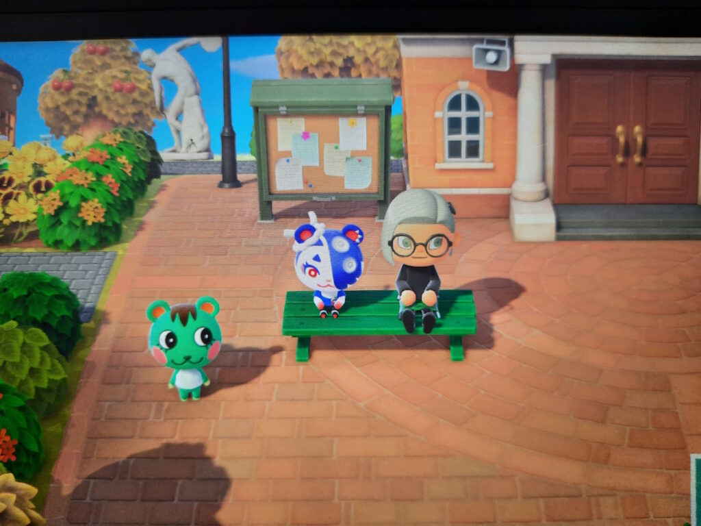 Ein Animal-Crossing-Screenshot, der meine Figur auf einer Bank neben einem blau-weißem Eichhörnchen. Ein weiteres, grünes Eichhörnchen macht neben der Bank Sport.