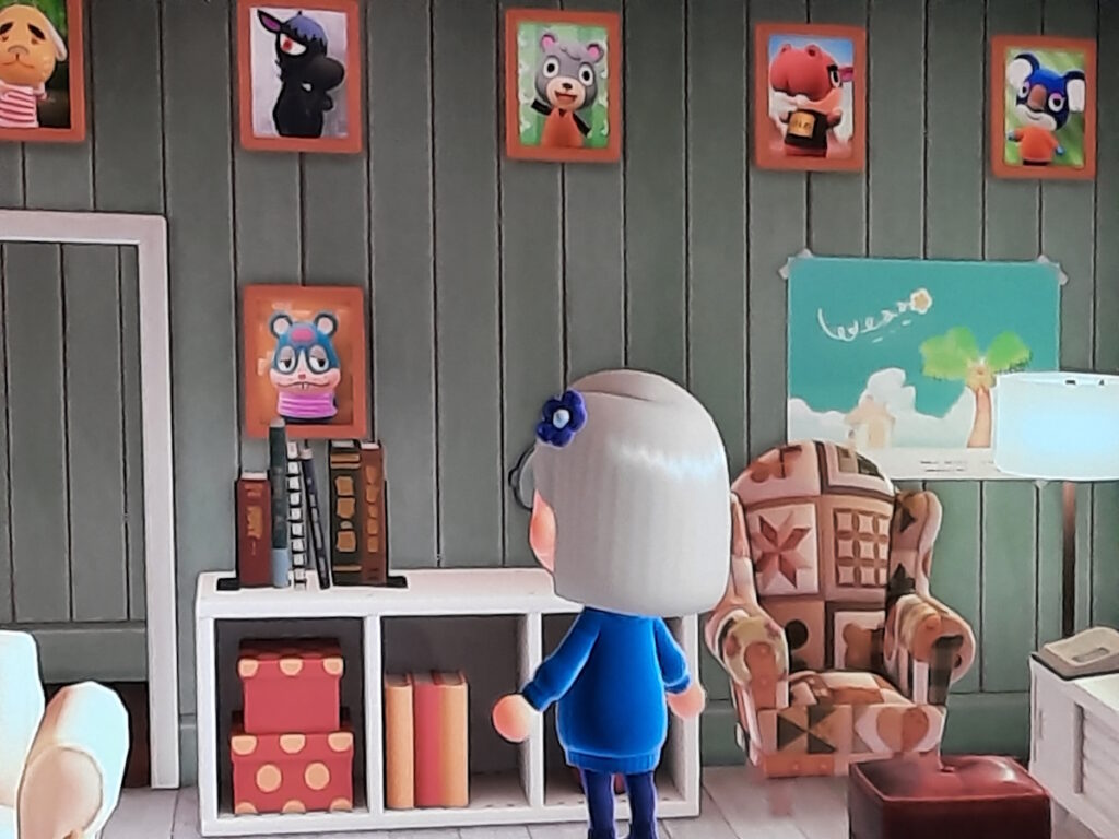 Meine Animal-Crossing-Figur steht mit dem Rücken zum Betrachter vor einer zartgrün getäfelten Wand. An der Wand hängen diverse Fotos von Animal-Crossing-Nachbarn. Eines davon ist von einem blauem Hamster mit mit einem breiten Mund. Unter dem Bild ist ein kleines Regal, auf dem ein paar Bücher stehen. Daneben steht ein gemütlicher Ohrensessel.
