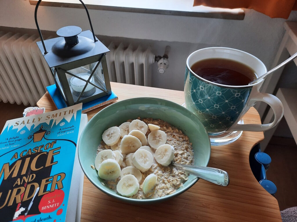 Eine zartgrüne Schale mit Porridge und Bananenscheiben zwischen der Taschenbuchausgabe von "A Case of Mice and Murder" und einer dampfenden Tasse mit Pfefferminztee.