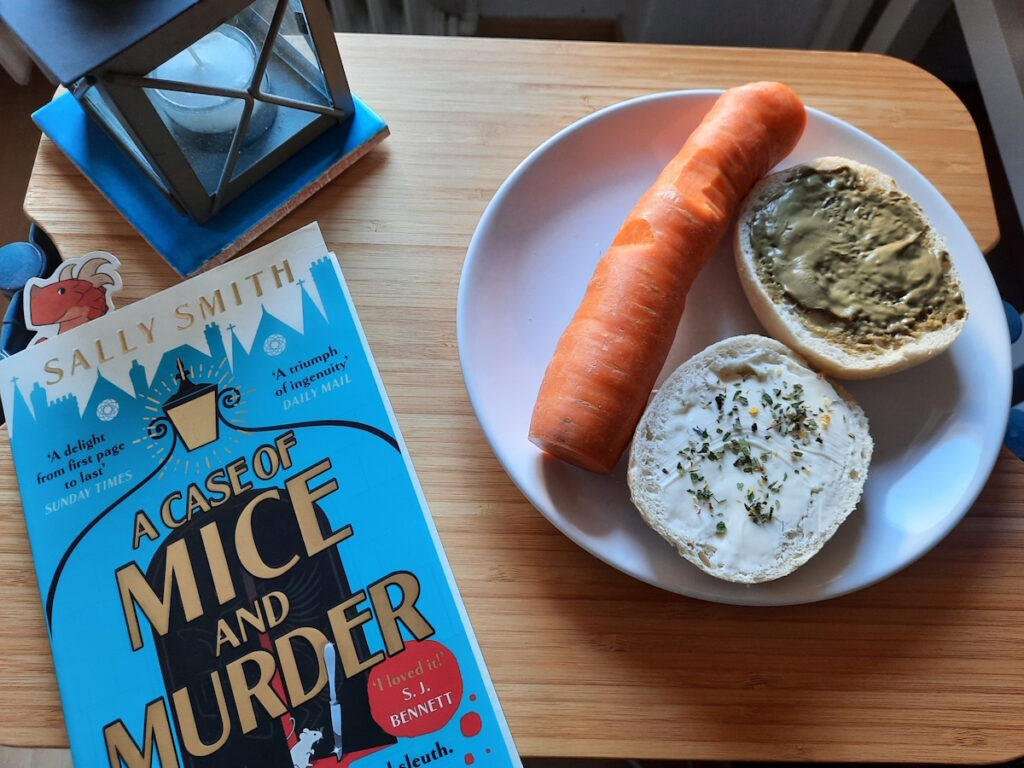 Ein kleiner Tisch auf dem rechts ein Teller mit einer riesigen Möhre und zwei Brötchenhälften steht. Die eine Hälfte ist mit Butter und Kräutersalz, die andere mit Pistaziencreme. Links davon liegt mein aktuelles Buch und dahinter steht eine kleine Sturmlaterne.