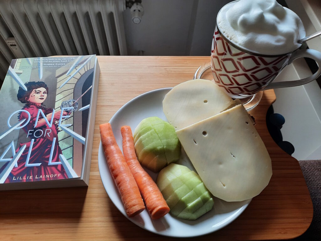 Ein Teller mit einer kleinen Kohlrabi, die in Stücke geschnitten wurde, zwei kleinen Möhren und einem halbiertem Käsebrot. Dahinter eine Tasse mit Milchkaffee, die von einer üppigen Milchschaumhaube gekrönt wird. Links davon die Taschenbuchausgabe von "One For All", dessen Cover eine junge Frau mit hochgesteckten dunklen Haaren, einem rotem Kleid und einem Florette in der Hand zeigt.