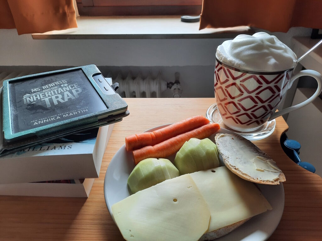 Ein Teller mit einem Käsebrot, einer kleinen, in Stücke geschnittenen Kohlrabi und zwei Möhren. Dahinter eine große Tasse mit Milchschaumhaube, in der ein Chai Latte ist. Links davon ein Buchstapel, auf dem ein angeschalteter eReader liegt. Der Bildschirm zeigt das Cover von "Ms. Renfield and the Inheritance Trap".