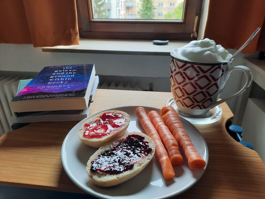 Ein kleiner Tisch auf dem im Vordergrund ein Teller mit zwei Brötchenhälften und drei sehr dünnen Möhren zu sehen ist. Die Brötchen sind einmal mit rotem und einmal mit schwarzem Johannisbeer-Fruchtaufstrich belegt. Im Hintergrund ist eine große Tasse Milchkaffee mit einer üppigen Milchschaumhaube zu sehen und auf der linken Seite ist ein kleiner Bücherstapel, dessen oberstes Buch "The Galaxy and the Ground Within" von Becky Chambers ist.