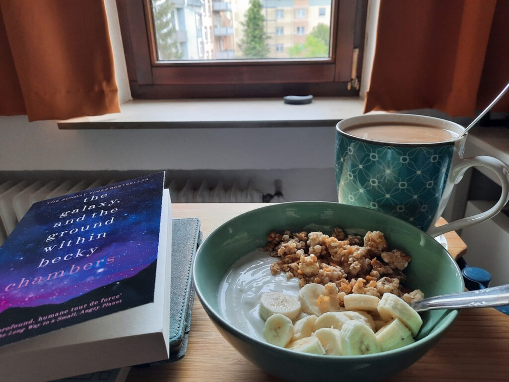 Ein kleiner Tisch mit einer Schale, die mit Skyr, Bananenscheiben und Haferknusperstückchen gefüllt ist. Dahinter steht eine große Tasse Tee und daneben liegt ein kleiner Buchstapel.