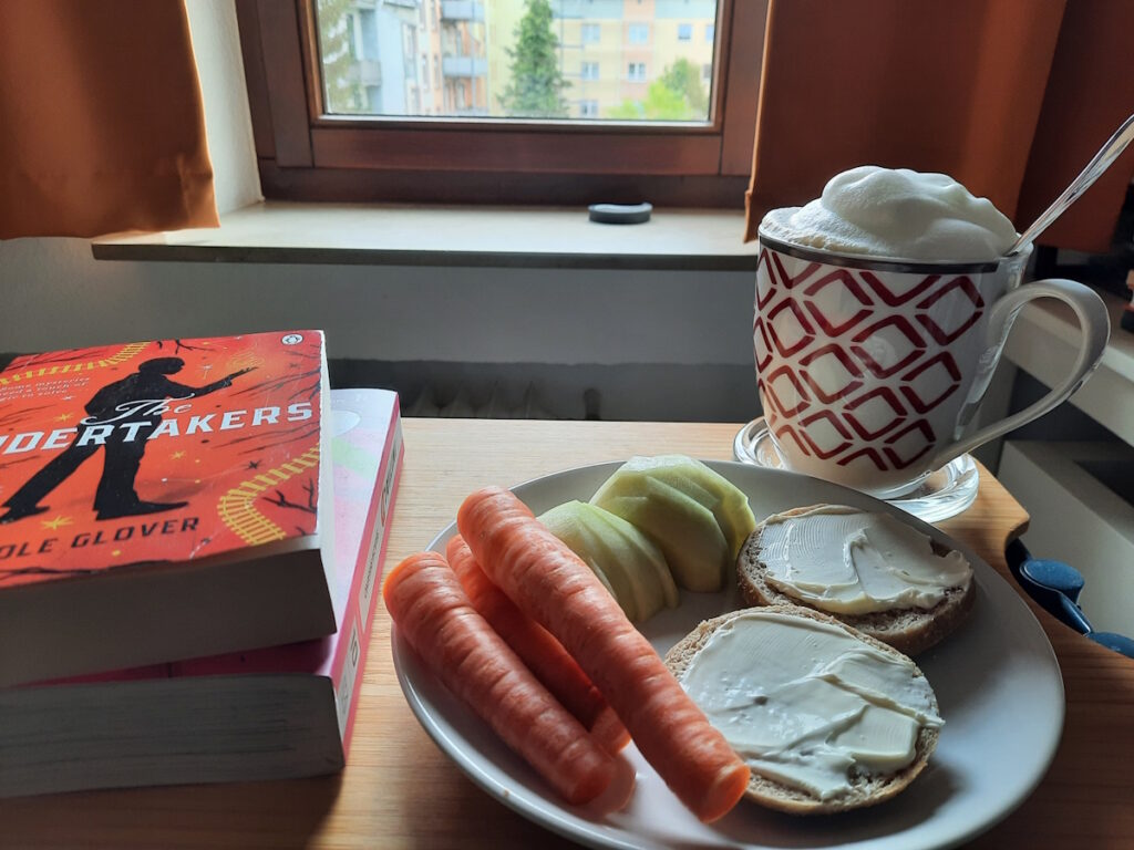 Ein Teller mit einer in Stücke geschnittenen kleinen Kohlrabi, drei kleinen Möhren und einem mit Frischkäse bestrichenem English Muffin. Dahinter eine große Tasse Milchkaffee, von dem über den Tassenrand hinweg nur eine üppige Milchschaumhaube zu sehen ist. Links davon liegen zwei Bücher aufeinander. Der Titel des oberen Buchs lautet "The Undertakers" von Nicole Glover.