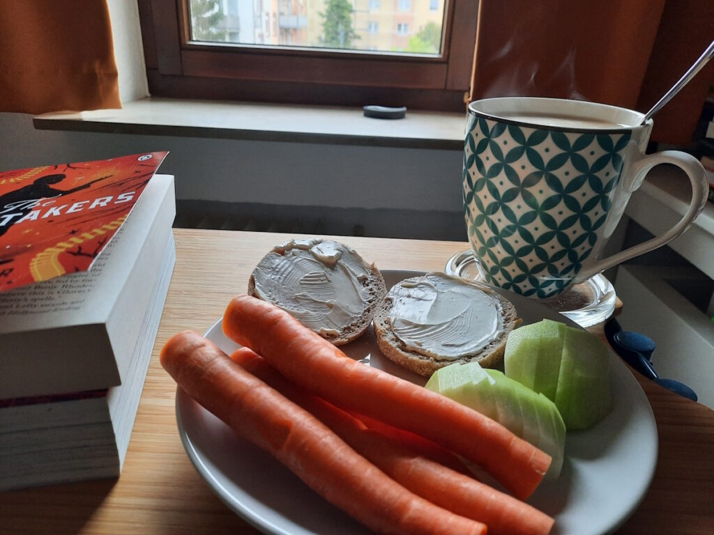 Ein Teller mit einer kleinen, in Stücke geschnittenen Kohlrabi, mehreren dünnen Möhren und einem English Muffin, der mit Frischkäse bestrichen wurde. Im Hintergrund dampft eine große Tasse mit Tee.