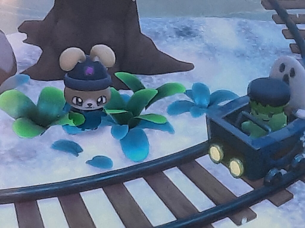 Meine Hello-Kitty-Island-Adventure-Figur steht vor einem Baum und schaut auf Schienen. Auf den Schienen nähert sich eine kleine Loren-Bahn mit einer grünen Figur, die einen "Frankensteins Monster"-Hut trägt und hinter der ein kleiner Geist zu sehen ist.