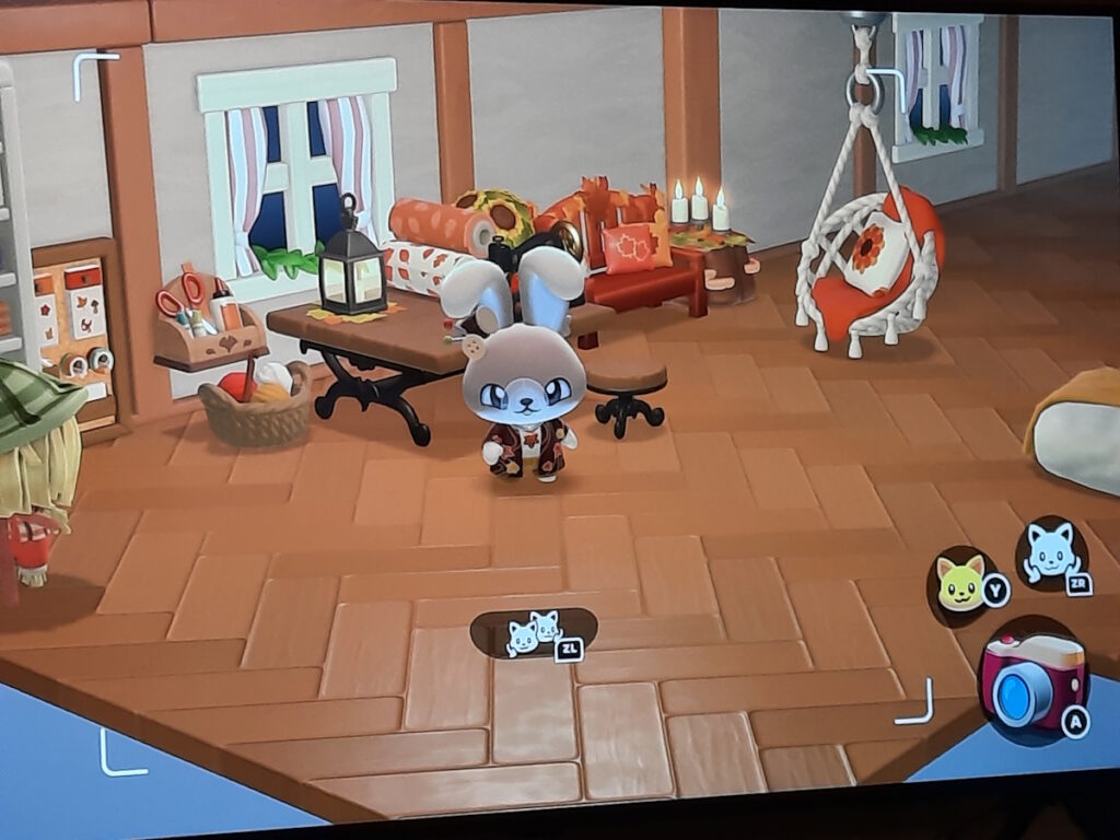 Ein Screenshot von "Hello Kitty Island Adventure". Mein Charakter, ein hellbrauner Hase, steht vor einem Tisch mit einer Nähmaschine und weiterem Nähzubehör, sowie einer Laterne, die mit Herbstblättern dekoriert ist. Im Hintergrund ist eine ebenfalls herbstlich dekorierte Gartenbank und ein gemütlicher Hängesessel mit einem kuscheligen Kissen zu sehen. Neben dem Tisch steht ein Korb mit Strickzeug.