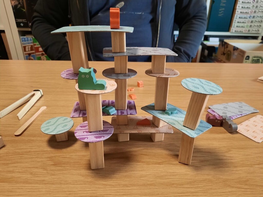 Ein Spiel, bei dem mehrere aufrecht stehende Hölzer mit Plattformen versehen wurden. Das Ganze sieht aus wie ein windschiefer Kratzturm für Katzen. Auf zwei verschiedenen Ebenen sitzen eine orange und eine grüne Holzkatze, außerdem lassen sich auf den verschiedenen Plattformen verschiedenes Katzenspielzeug erahnen.