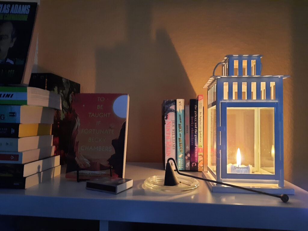 Eine brennende Kerze in einer ca. 30 cm hohen, weißen Sturmlaterne. Hinter der Kerze stehen einige Bücher zwischen  zwei Buchstützen. Links davon ist ein Buch mit dem Cover zum Betrachter aufgestellt, es handelt sich um den Titel To Be Taught, If Fortunate von Becky Chambers. Am linken Bildrand ist ein Stapel Bücher zu erahnen.