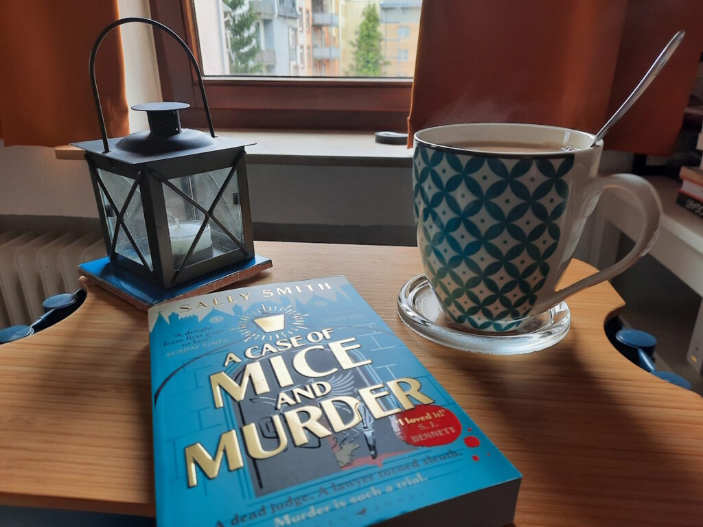 Ein kleiner Tisch mit einer großen, dampfenden Tasse Tee im Hintergrund und der Taschenbuchausgabe von "A Case of Mice and Murder" im Vordergrund. Das hellblaue Cover zeigt eine stilisierte altmodische Bogentür in dessen offenem Türflügel eine weiße Maus vor einem Messer und einer Blutlache sitzt. Darüber steht in großen, goldenen Buchstaben der Titel.