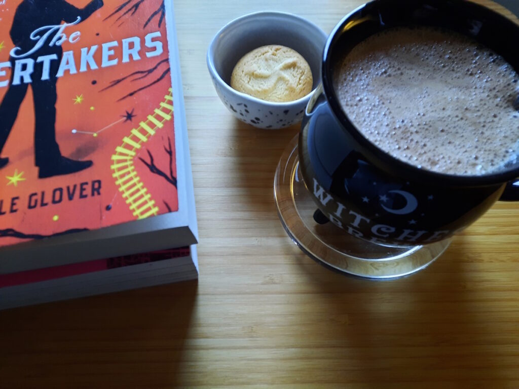 Vorne rechts steht eine Tasse, die wie ein Hexenkessel geformt ist und in der sich Heiße Schokolade befindet. Links daneben ist eine kleine Schale mit zwei Zitronenkeksen und daneben liegt das Buch "The Undertakers".