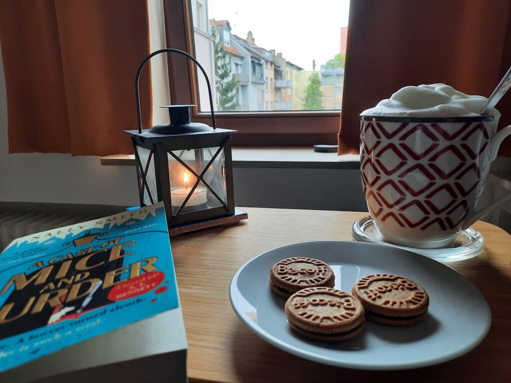 Im Vordergrund rechts eine Untertasse mit drei Keksen und links mein aktuelles Buch. Im Hintergrund rechts eine große Tasse Chai mit einer Milchschaumhaube und links davon eine kleine brennende Sturmlaterne.