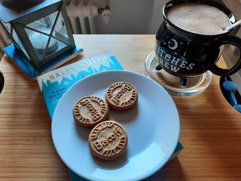 Eine Untertasse mit drei Keksen auf der Taschenbuchausgabe von A Case of Mice and Murder und dahinter eine schwarze Tasse mit der Aufschrift Witches Brew. Die Tasse ist mit schaumiger Heißer Schokolade gefüllt.