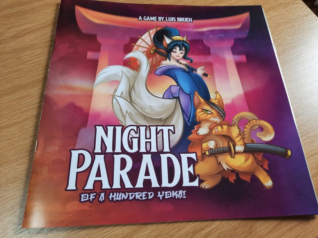Die Schachtel des Spiels Night Parade von Luis Brueh. Das Cover zeigt eine japanisch anmutende Figur mit mehreren weißen Fuchsschwänzen vor einem weißen Torii steht. Vor dieser Person steht eine Katze auf ihren Hinterbeinen mit einem Schwert in der Pfote.