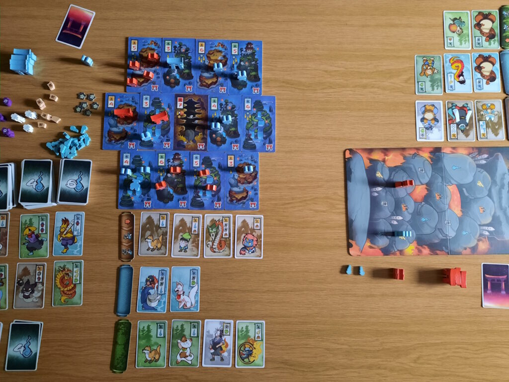 Überblick über den Spielaufbau von Night Parade. In der Mitte ist ein dunkelblaues Spielbrett aus mehreren Teilen zusammengesetzt. Darunter liegen mehrere aufgedeckte Karten mit verschiedenen Yokai-Darstellungen und kleinen Symbolen. Außerdem sind noch viele kleine Holzteile und Kartenstapel zu sehen.