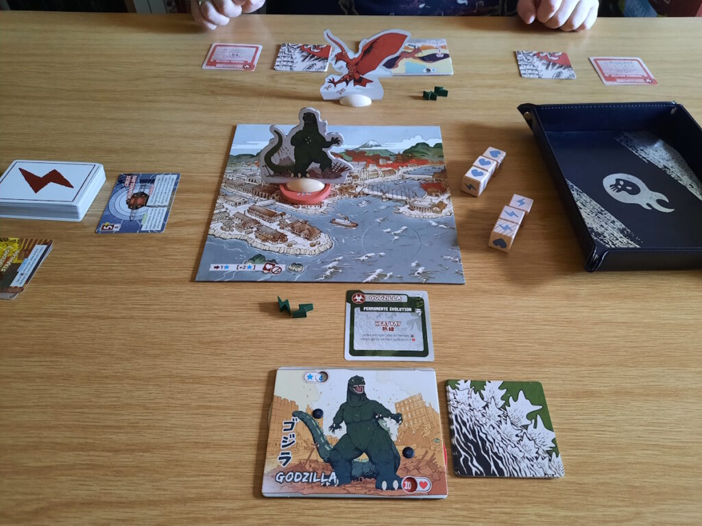 Ein Spielbrett, dass Tokio und die Tokioer Bucht andeutet und auf dem eine Godzilla-Figur steht. im Hintergrund ist Rodan zu erahnen, der von meinem Mann gespielt wurde. Im Vordergrund liegen mehrere Karten, die die möglichen Aktionen beschreiben.