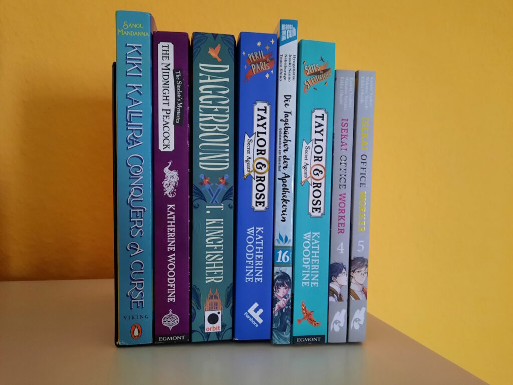 Einige Bücher und Manga zwischen zwei Buchstützen. Die Titel lauten von links nach rechts: Kiki Kallira Conquers a Curse, The Midnight Peacock, Daggerbound, Peril in Paris, Die Tagebücher der Apothekerin 16, Spies in St. Petersburg, Isekai Office Worker 4+5