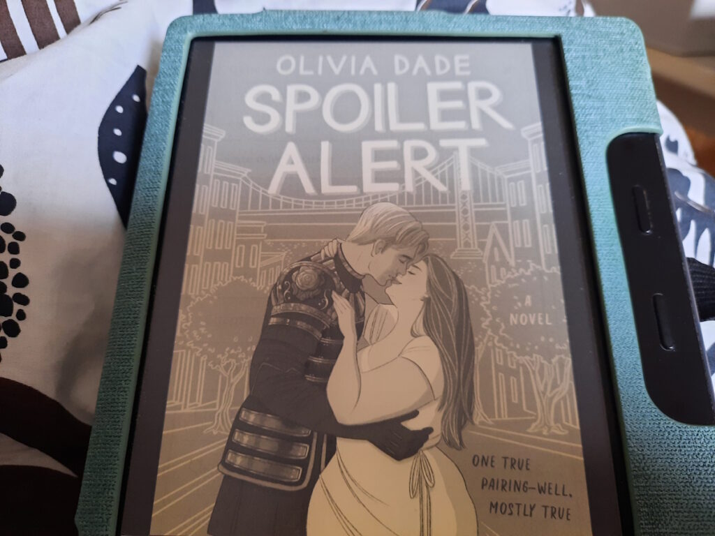 Ein angeschalteter eReader, dessen Bildschirm das Cover von "Spoiler Alert" von Olivia Dade zeigt. Der große, blonde Protagonist trägt eine Uniform und beugt sich runter zu der kurvigen Protagonistin, die eine Art klassisches griechisches Gewandt trägt und deren offene Harre in einer üppigen Mähne über ihren Rücken fallen.