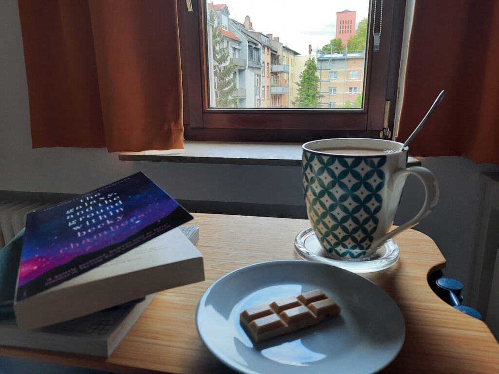 Eine Untertasse mit zwei kleinen Riegeln Karamellschokolade. Dahinter eine große Tasse mit Milchtee und daneben ein kleiner Stapel mit Büchern.