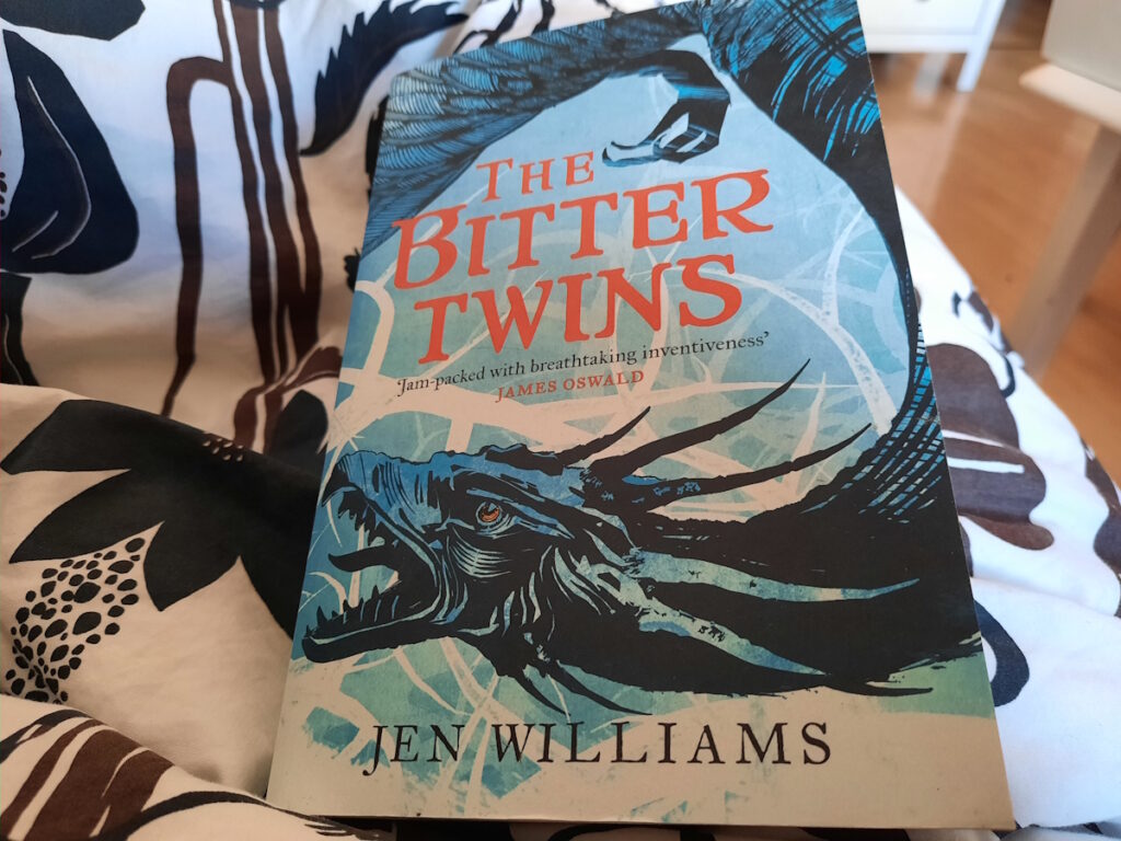 Die Taschenbuchausgabe von "The Bitter Twins" auf einer gemusterten Decke. Das Cover ist in hellblau gehalten und zeigt einen dunklen blaugrauen Drachen, der sich einmal um den in roter Schrift gehaltenen Titel windet.