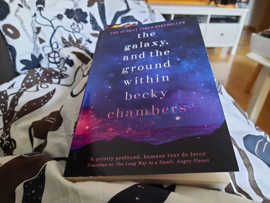 Ein dunkelblaues Buch auf einer Decke mit braun-schwarzen Blumen auf weißem Hintergrund. Das Buch trägt den Titel "The Galaxy and the Ground Within" von Becky Chambers und das Cover deutet am unteren Rand schwarze Landmasse und einen violetten Ozean an, der dunkelblaue Himmel ist von Sternen durchzogen.