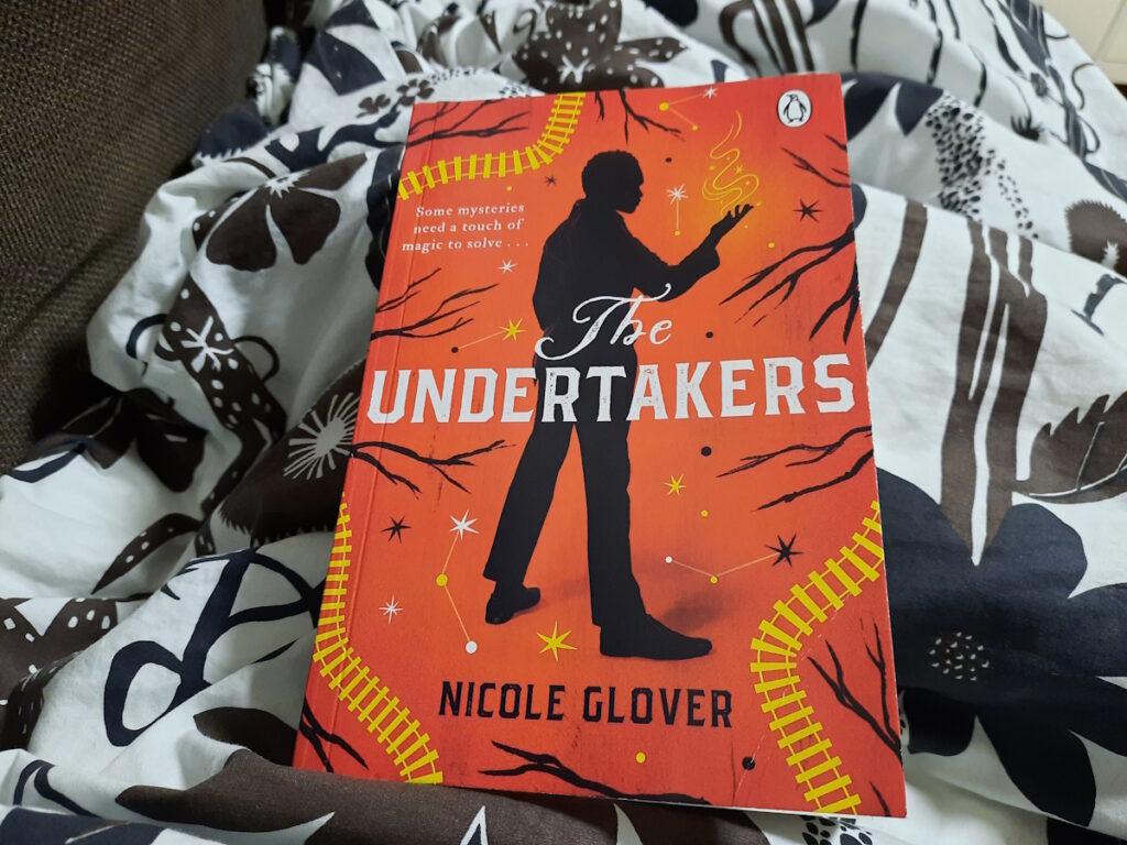 Die Taschenbuchausgabe von "The Undertakers" von Nicole Glover. Das orange Cover zeigt die Silhouette eines Mannes von schräg hinten. Er hat die rechte Hand erhoben und es scheint ein Leuchten von ihr auszugehen.
