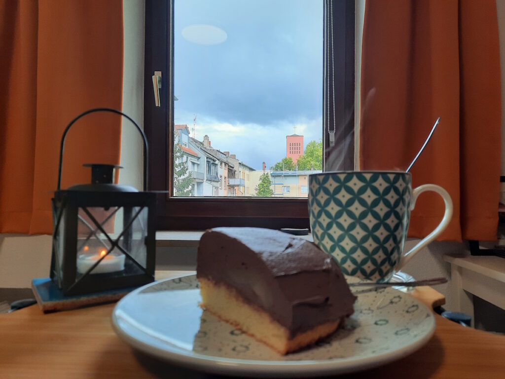 Im Vordergrund ein großes Stück Schokoladen-Birnen-Torte, dahinter eine große dampfende Tasse Tee. Links davon eine kleine Sturmlaterne mit einer winzigen Flamme und im Hintergrund ein Fenster, durch den ein Himmel mit großen grauen Wolken zu erahnen ist.