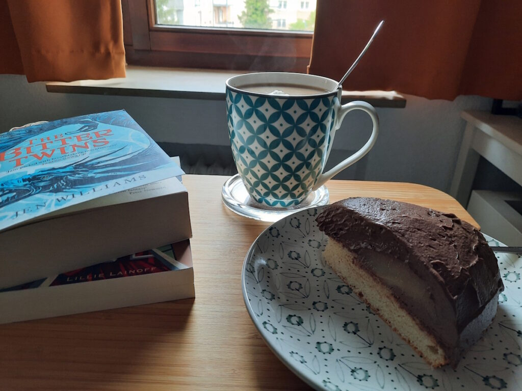 Rechts vorne steht ein Teller mit einer Schokoladen-Birnen-Torte mit einem hellen Bisquitboden. Dahinter eine große dampfende Tasse mit Tee und links davon ein kleiner Stapel aus zwei Taschenbüchern. Der oberste Titel lautet "The Bitter Twins".